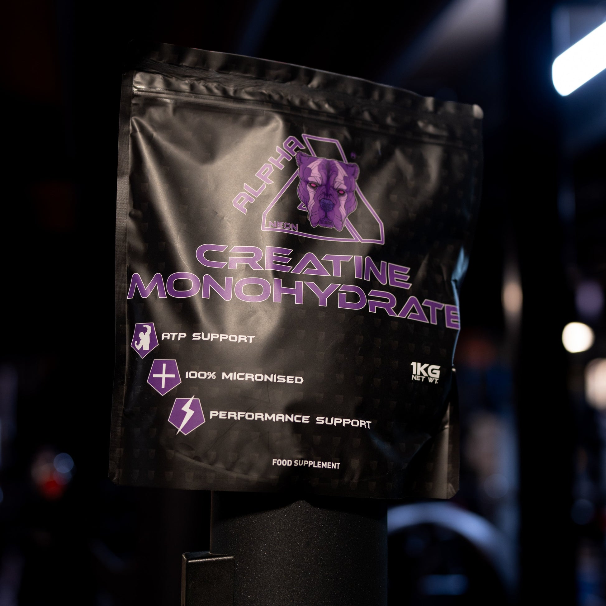 A black stand-up pouch of Creatine Monohydrate with purple text and logo, photographed on a dark gym floor with blurred equipment and lighting in the background.