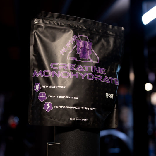 A black stand-up pouch of Creatine Monohydrate with purple text and logo, photographed on a dark gym floor with blurred equipment and lighting in the background.