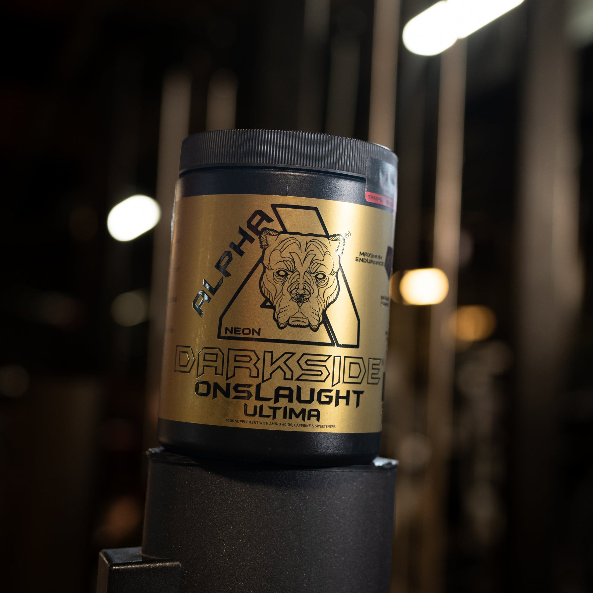 A black tub of Alpha Neon Darkside Onslaught Ultima supplement powder with a metallic gold label, set against a dark gym background with blurred lights.