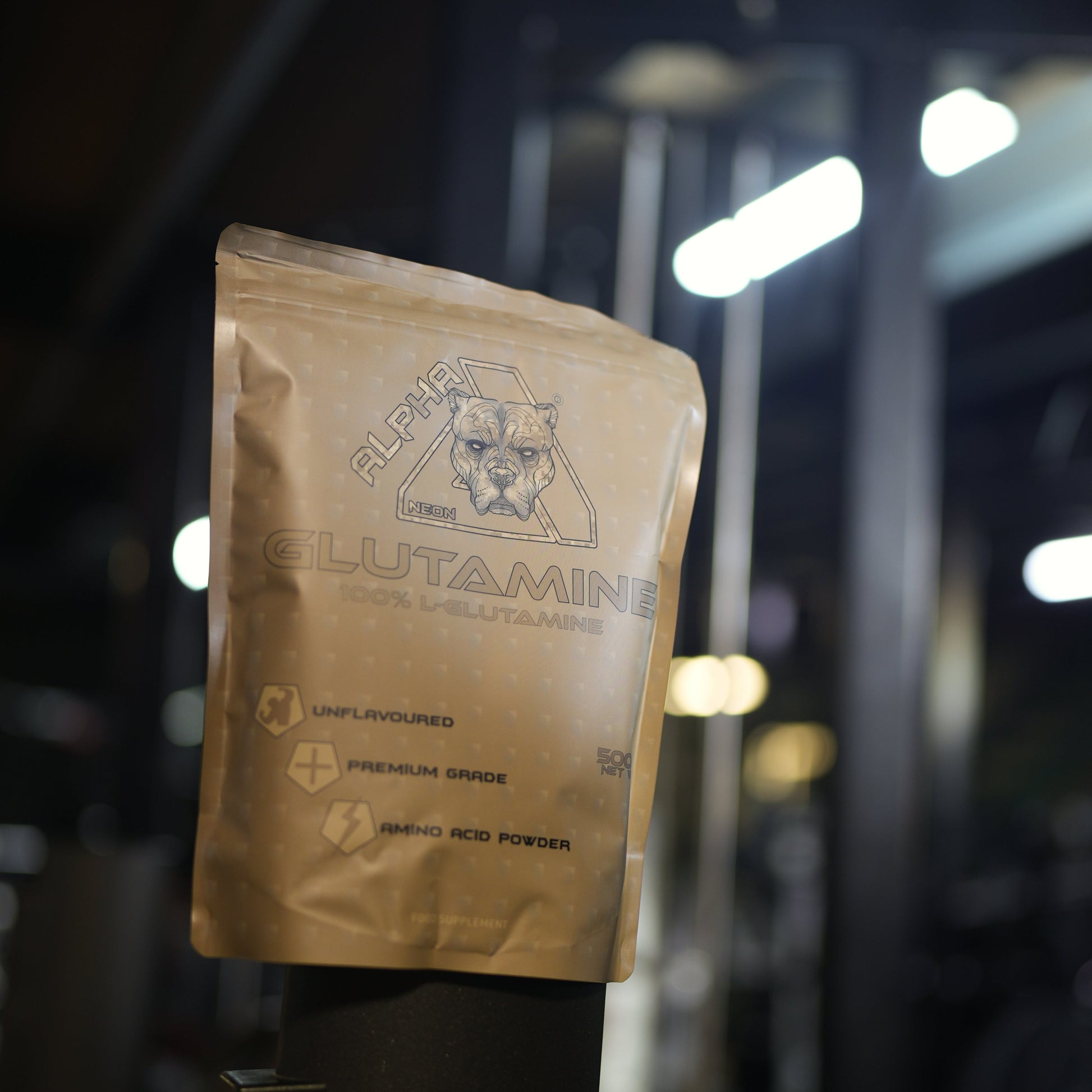 A stand-up pouch of Alpha Neon Glutamine Unflavoured powder, displayed on a black stand in a gym setting with blurred exercise equipment and lights in the background.