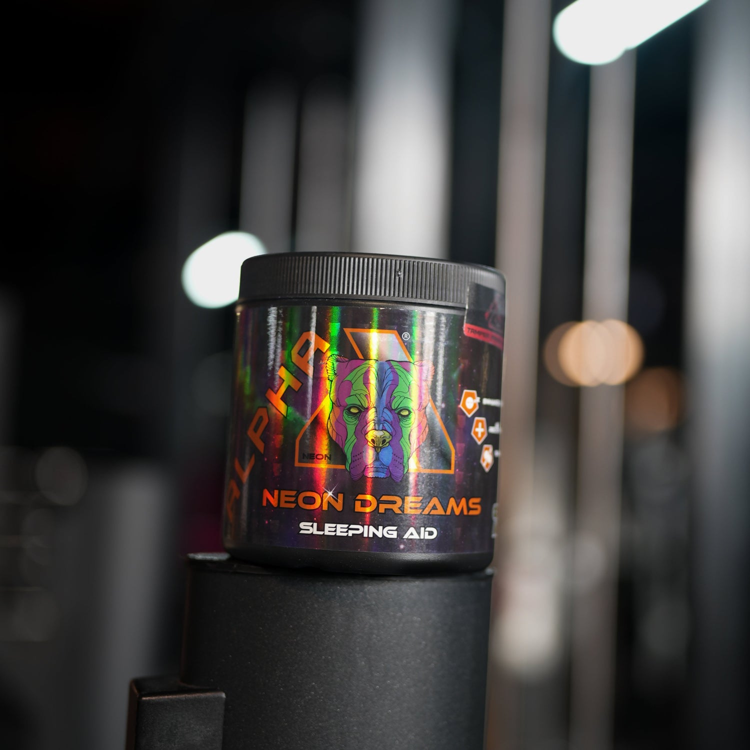 A black cylindrical container labeled 'NEON DREAMS SLEEPING AID' with a ribbed lid, shown resting on a black stand in a dimly lit gym environment. The label features a colorful neon pattern with orange, green, and pink text.