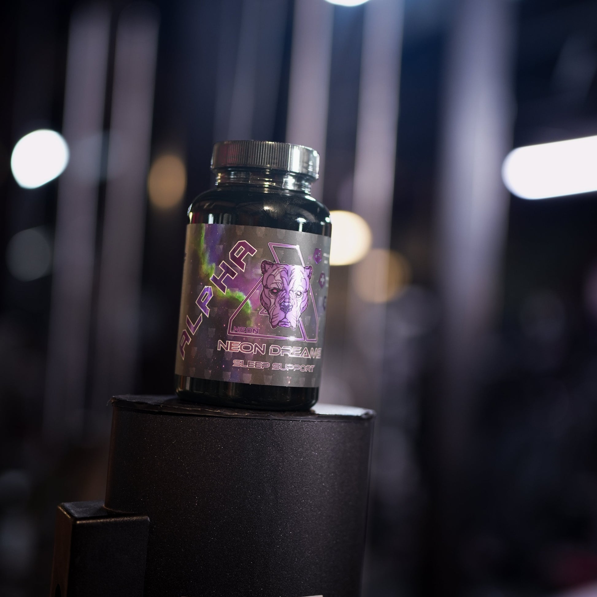 A small black supplement bottle with a metallic purple and pink label reading 'NEON DREAMS' sits on top of a black display stand in a dimly lit gym environment with blurred lights in the background.