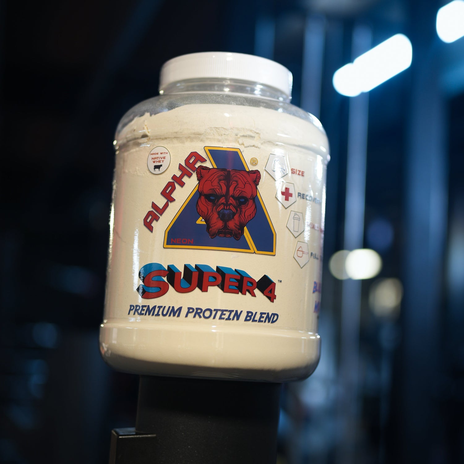 A large tub of Alpha Neon Super 4 Premium Protein Blend powder sits on a black stand in a gym setting with blurred exercise equipment and bright overhead lights in the background.
