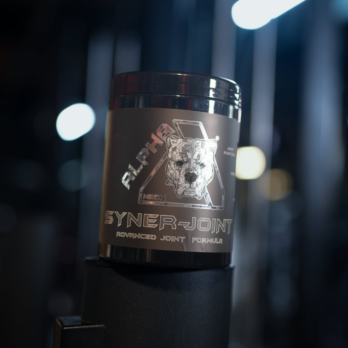 A black tub of Alpha Neon SynerJoint Advanced Joint Formula powder, featuring a stylized dog logo on the label, set against a dark, blurred gym background with bright overhead lights.