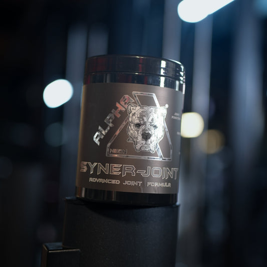 A black tub of Alpha Neon SynerJoint Advanced Joint Formula powder, featuring a stylized dog logo on the label, set against a dark, blurred gym background with bright overhead lights.